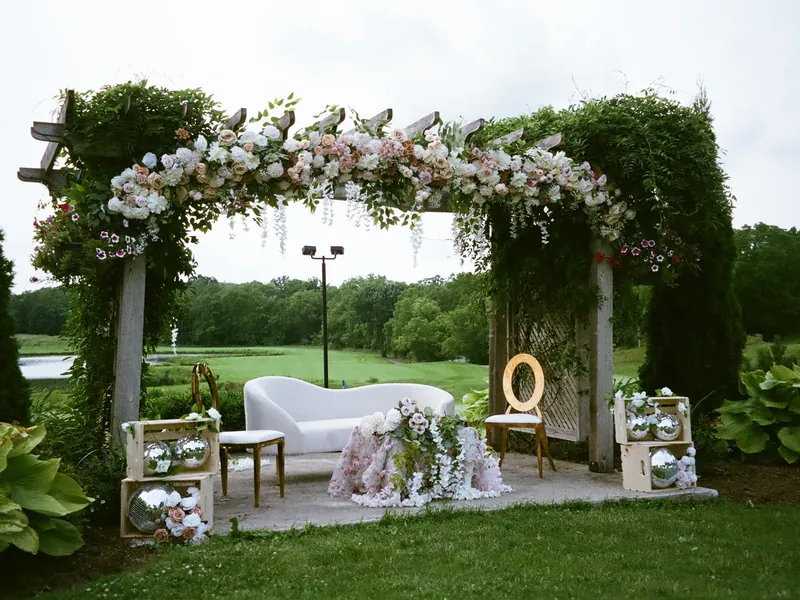 Outdoor wedding stage under a floral pergola with a white loveseat and disco balls on a golf course lawn